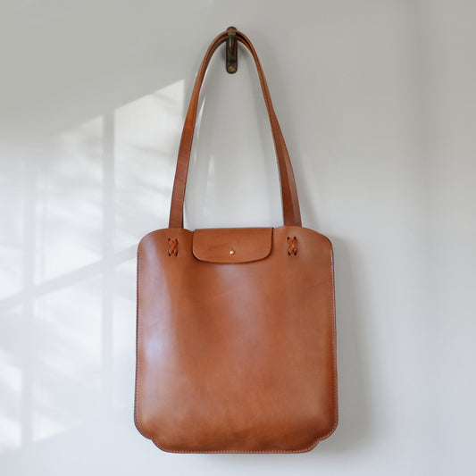 Scalloped Slim tote bag product image. Caramel color leather bag hanging on a hook against a white wall.
