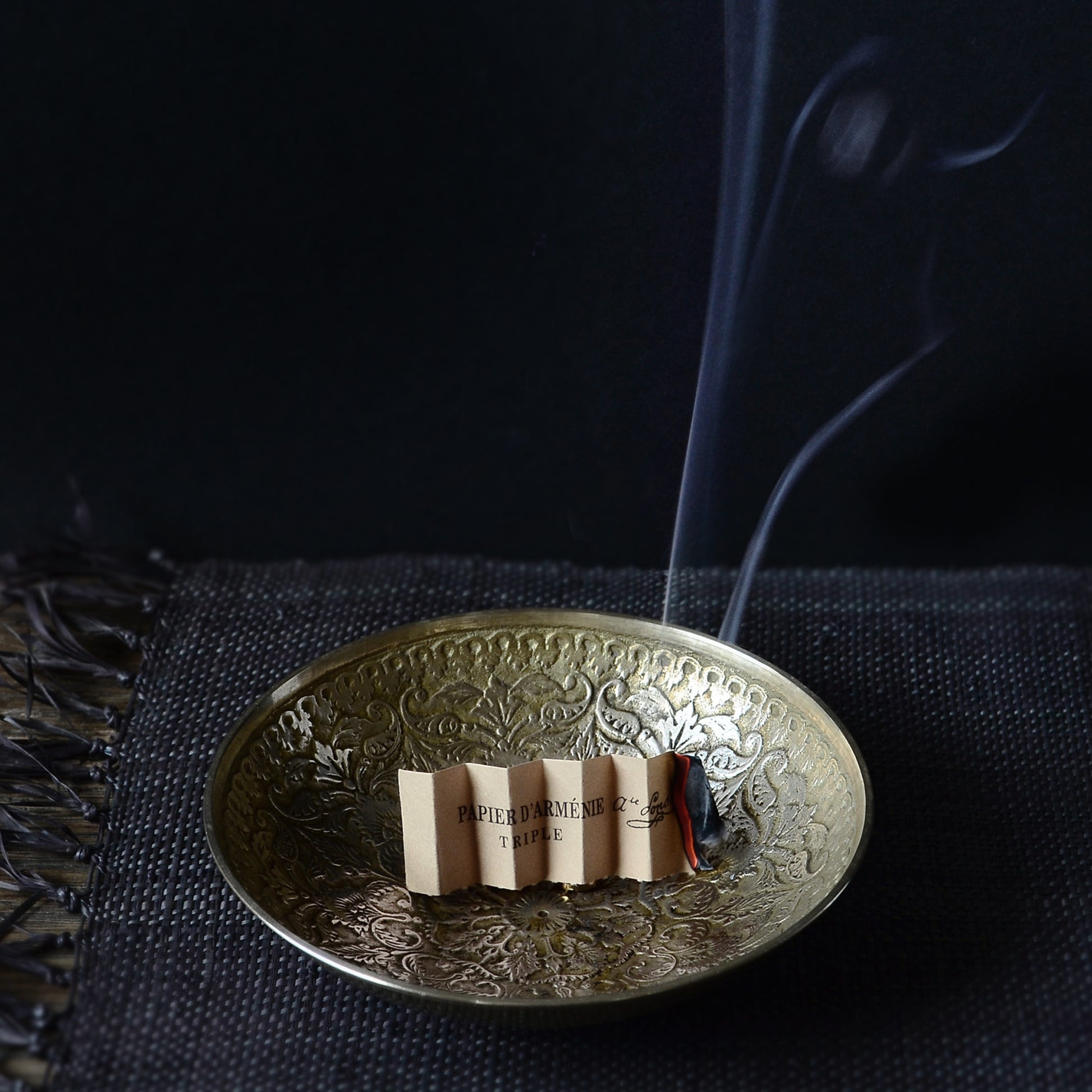 An accordion folded piece of Papier d'Armenie incense paper burning in a shallow brass bowl.