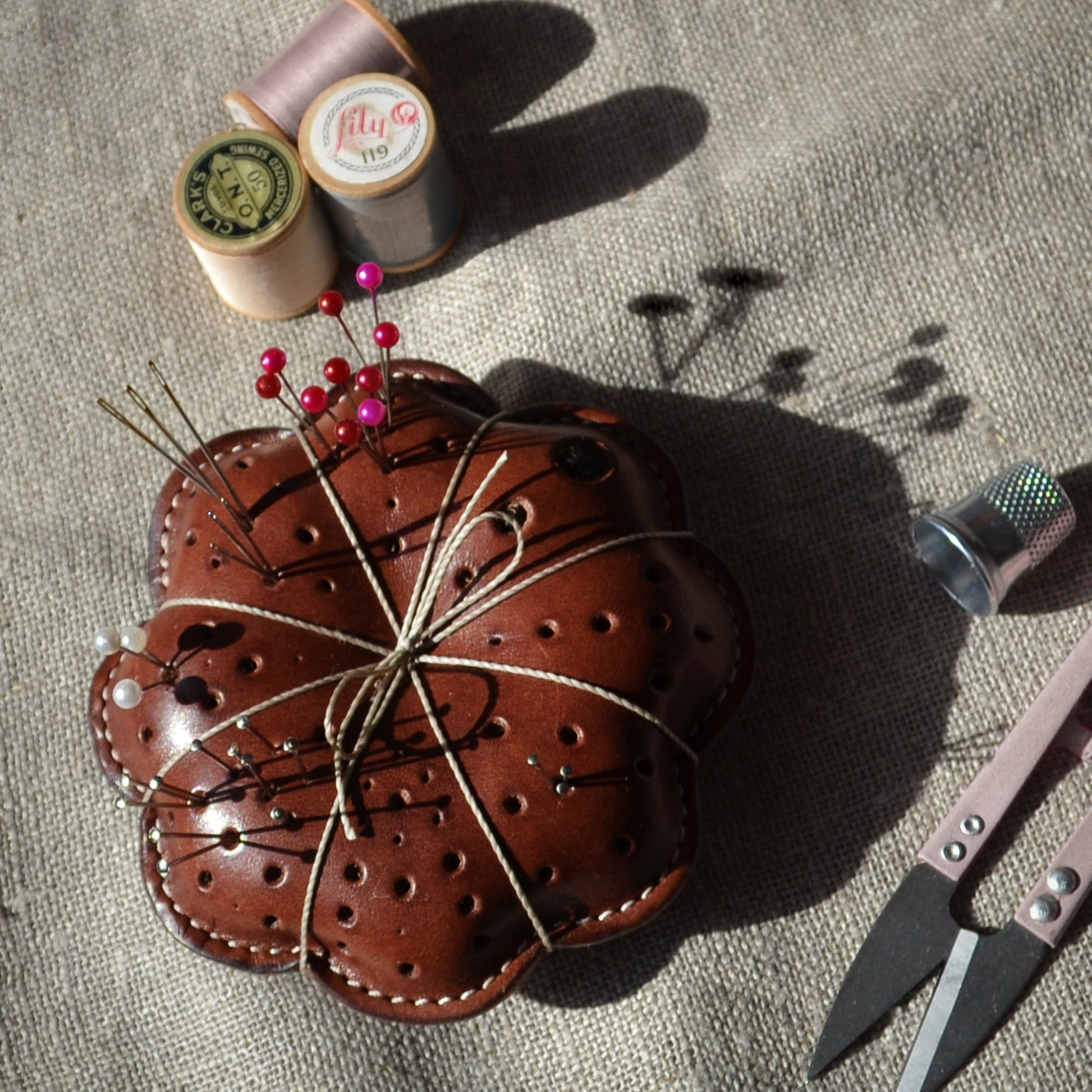Leather thread-wrapped pillow pin cushion with some pins and needles in it. Spools of thread, a thimble, and thread clippers sitting next to it.