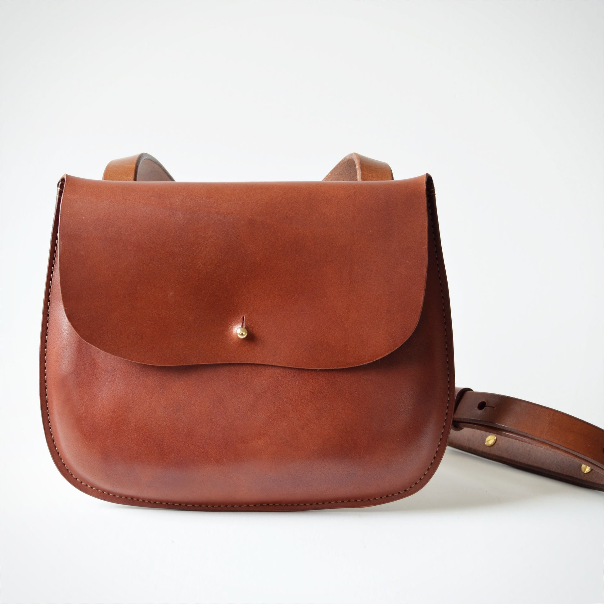 A medium brown color crossbody bag sits on a white surface with the strap looped to the side.