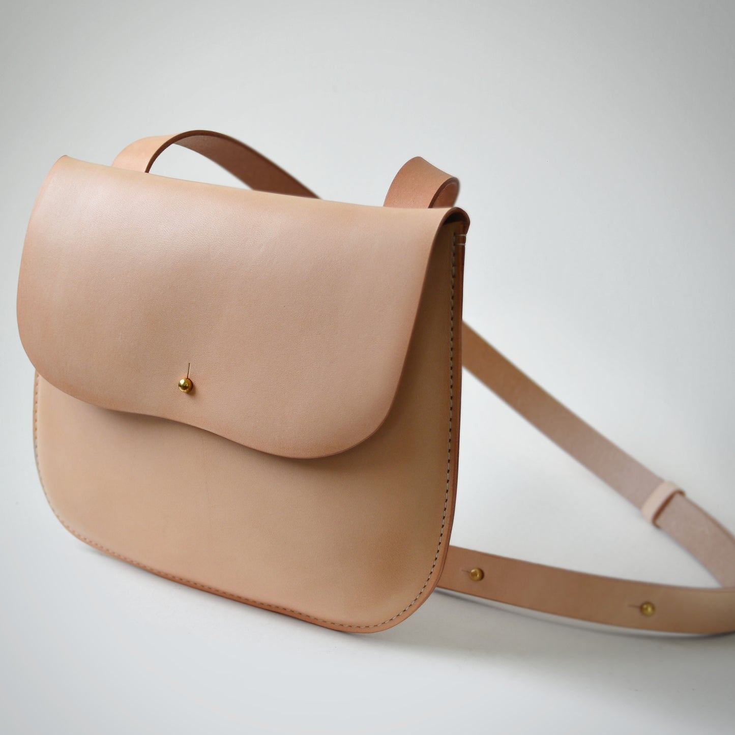 A natural color leather bag stands on a white background with the strap  looping off to the right.