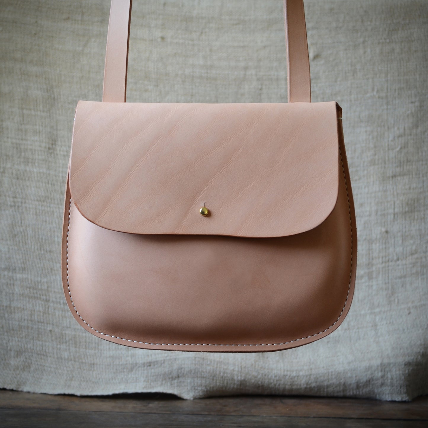 A natural color crossbody bag hangs in front of a woven  tan cloth