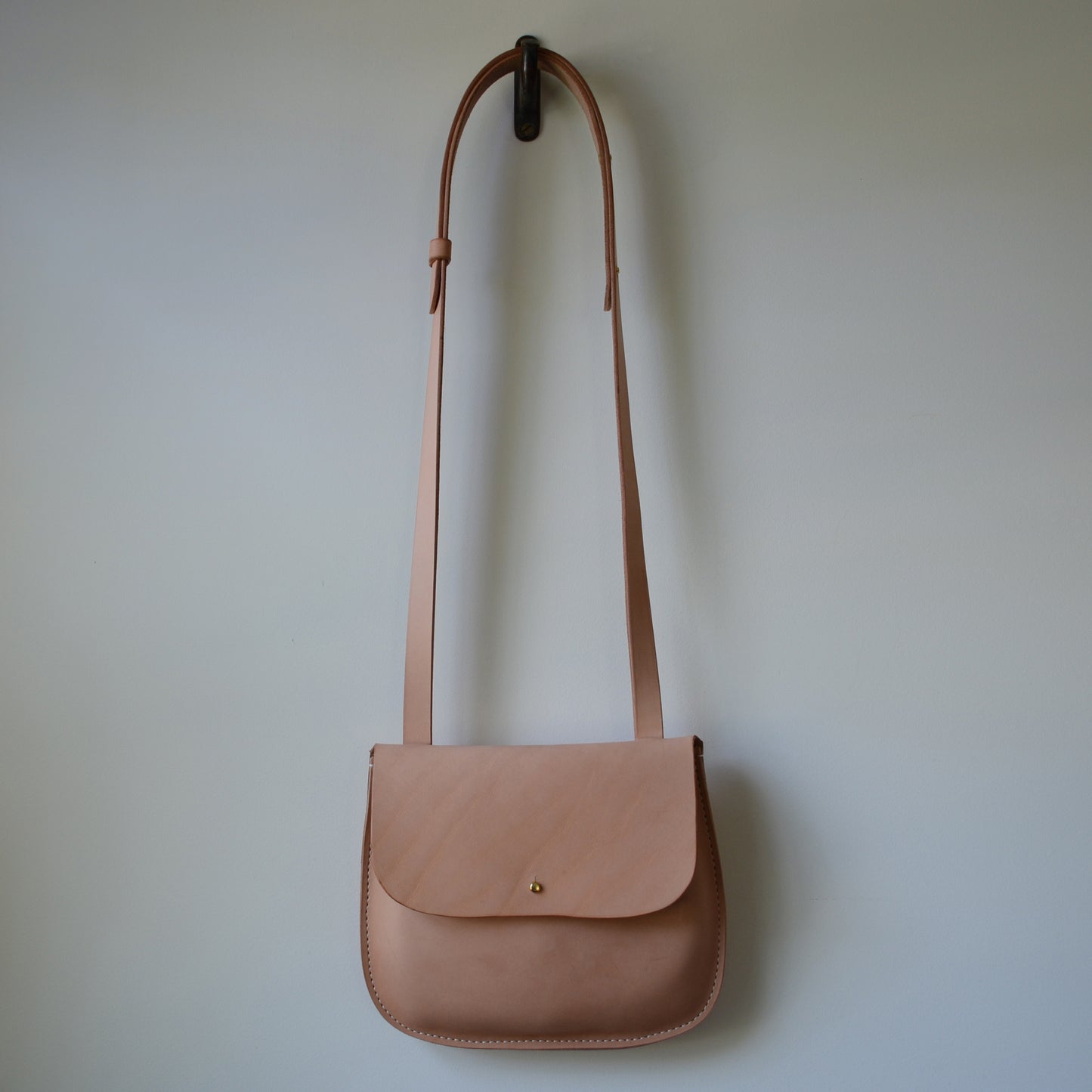 A natural color crossbody  bag hangs on a hook  on a white wall.