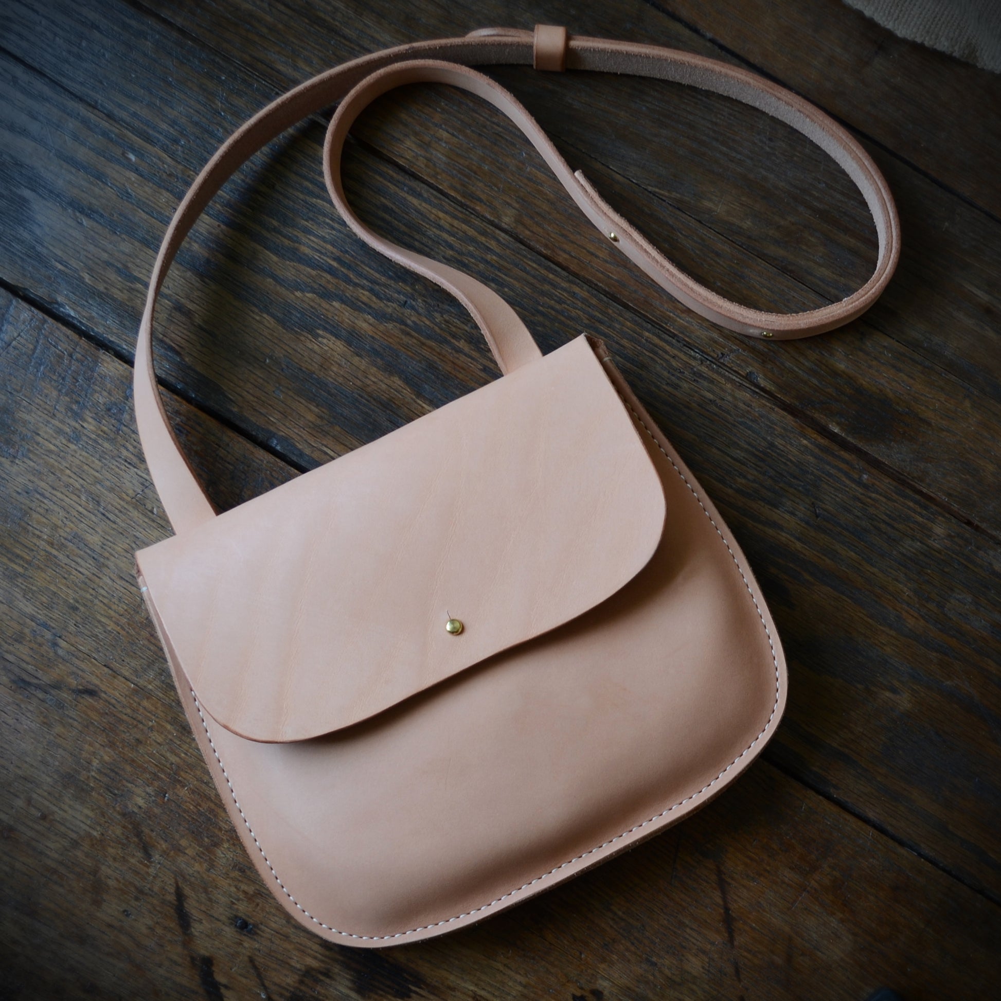 A natural color leather crossbody bag lays on a dark plank wood surface with the strap curling off to the side.