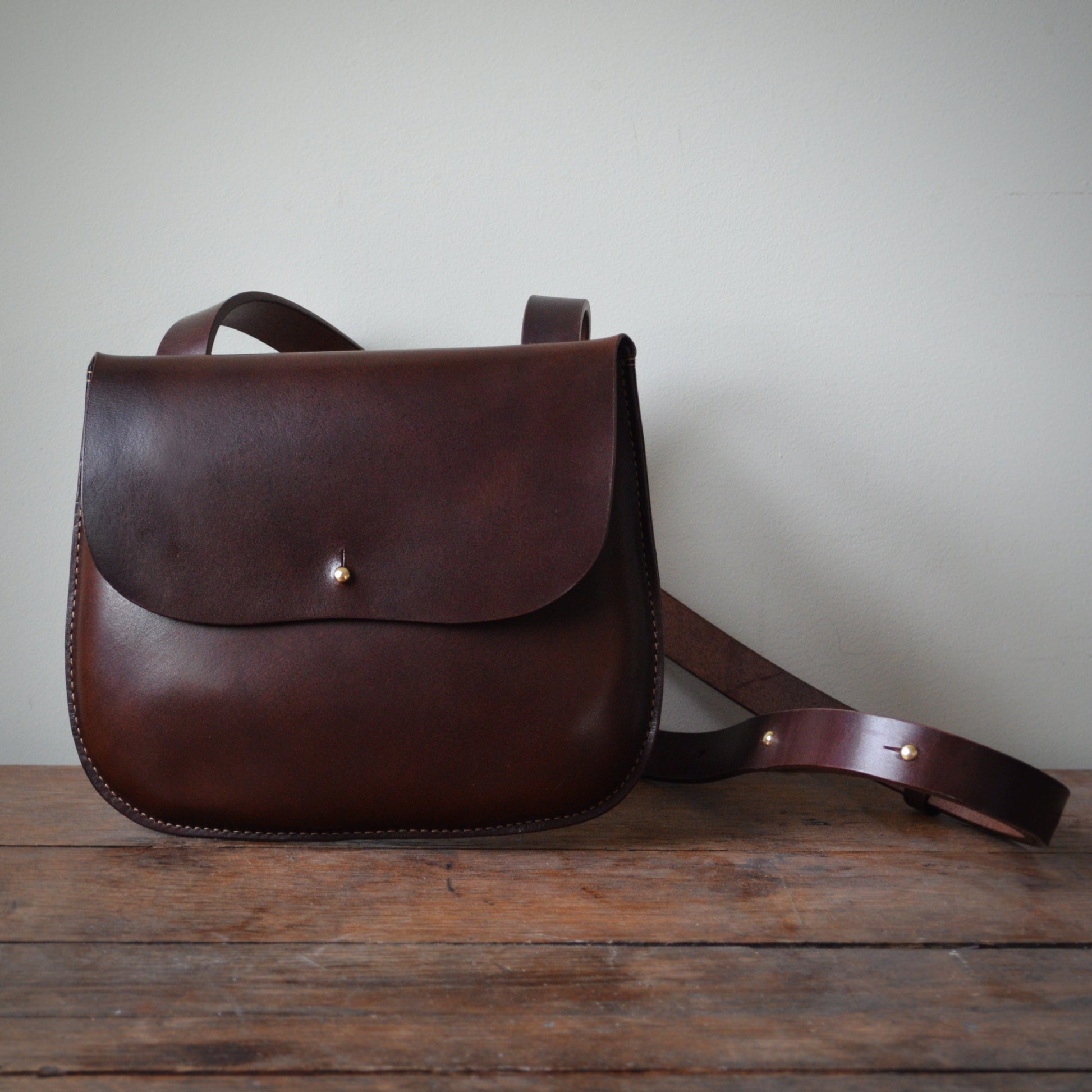 A dark brown crossbody bag sits on stands on a wood plank surface against a wall.