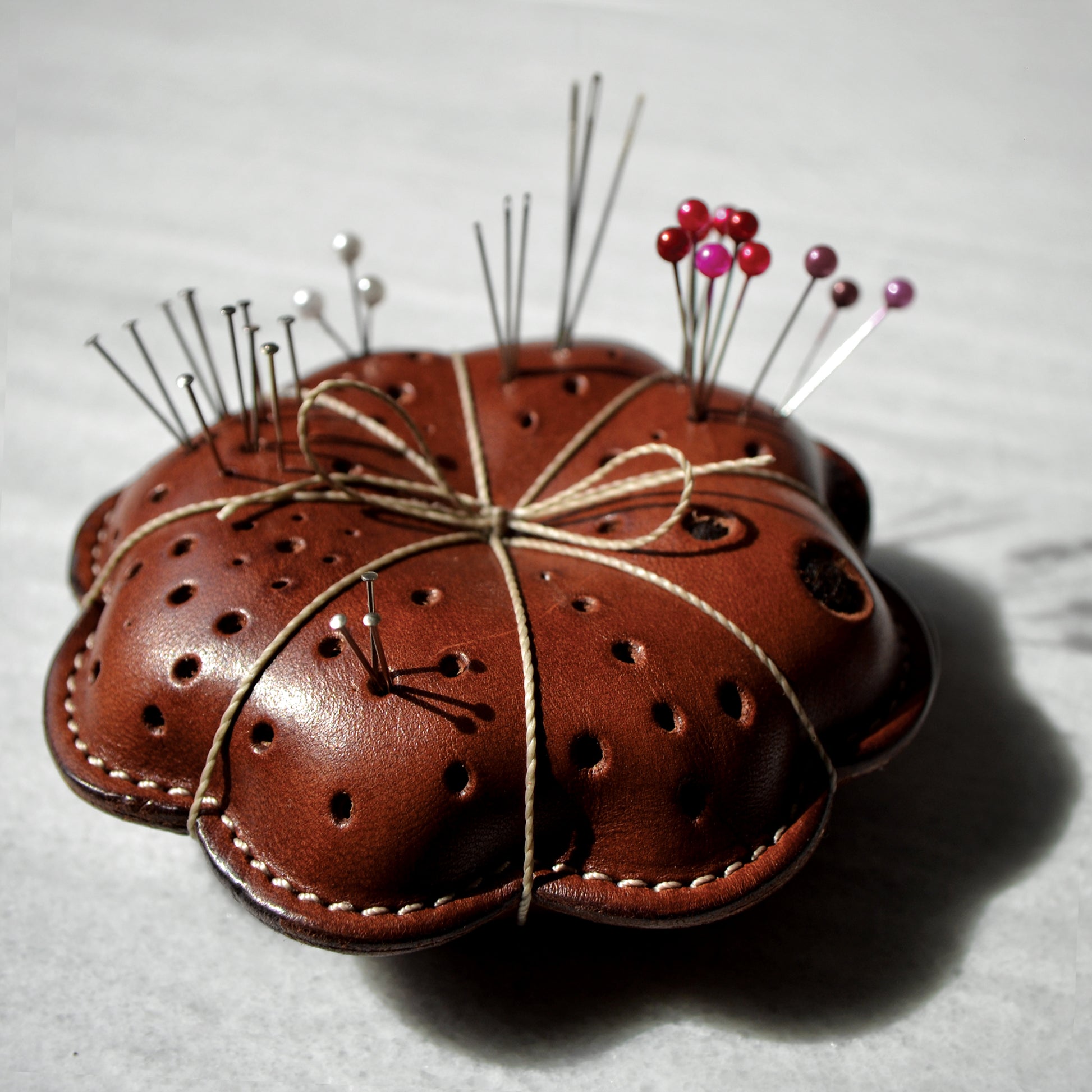 Leather thread-wrapped pillow pin cushion with some pins and needles in it.
